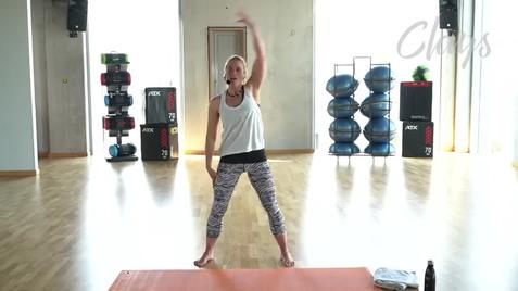 Frau in Sportkleidung mit hochgestrecktem Arm in einem Fitnessraum mit Trainingsgeräten hinter ihr.