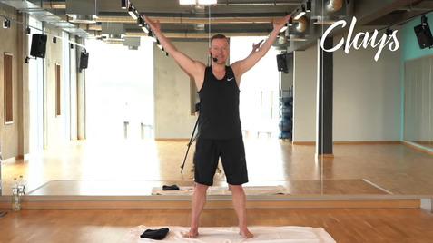 Mann mit erhobenen Armen steht auf einer Trainingsmatte in einem Fitnessraum mit Spiegeln.