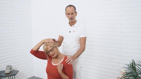 Ein Mann steht hinter einer sitzenden Frau und zeigt eine Dehnübung für den Nacken in einem modernen Raum.