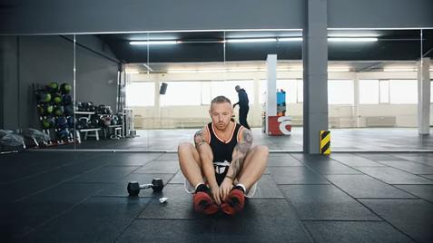 Mann im Fitnessstudio sitzt auf dem Boden und bindet seine Sportschuhe, im Hintergrund weitere Trainierende und Geräte.
