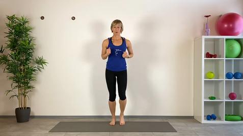 Frau in blauer Sportoberbekleidung steht barfuß auf einer Matte, umgeben von Fitnessgeräten und Pflanzen.