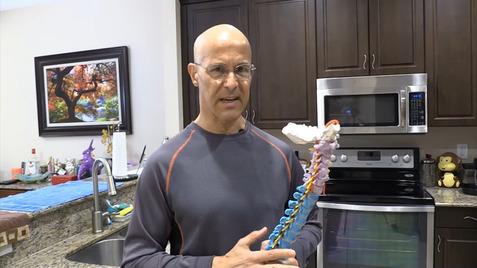 Mann in Sportkleidung hält ein anatomisches Modell der Wirbelsäule in einer modernen Küche.