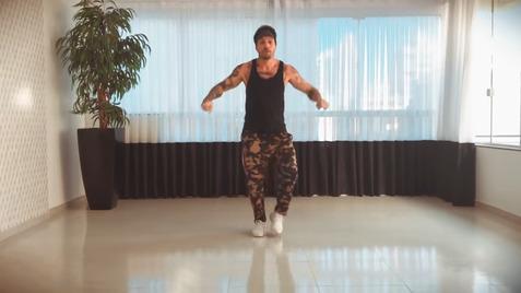 Mann tanzt in einem leeren Raum, trägt ein schwarzes Tanktop und camouflierte Trainingshosen, mit einer Pflanze im Hintergrund.