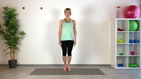 Frau steht auf einer Yogamatte in einem Fitnessraum mit Pflanzen und Sportgeräten im Hintergrund.