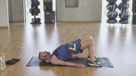 Mann in Sportkleidung führt eine Brückenübung auf einer Matratze im Fitnessstudio aus. Wasserflasche und Handtuch daneben.