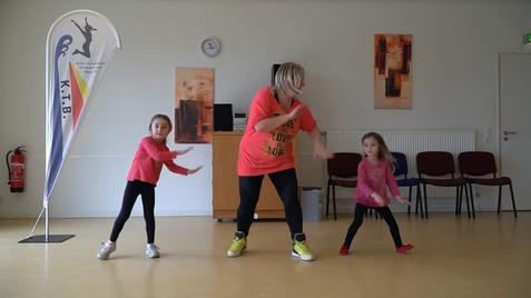 Tanztraining mit zwei Mädchen und einer Trainerin in einem Raum mit einer Fahne im Hintergrund.