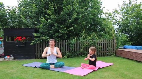 Frau und Kind praktizieren Yoga im Freien auf Matten, umgeben von Grün und einem Holzzaun im Hintergrund.