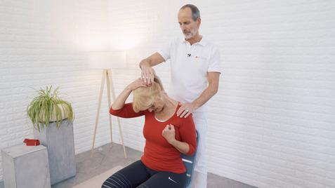 Ein Mann führt eine Nackenmassage bei einer sitzenden Frau in einem hellen Raum mit Pflanzen und einer Lampe durch.