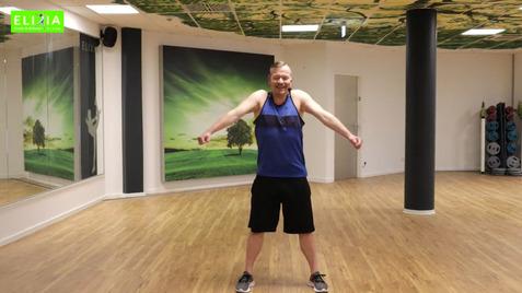 Mann in Sportbekleidung steht mit ausgestreckten Armen in einem Fitnessraum mit Spiegeln und grüner Wandgestaltung.