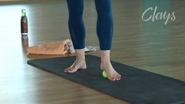 Füße stehen barfuß auf einer Yogamatte, während ein grüner Ball und Handtuch daneben liegen. Eine Flasche Wasser im Hintergrund.