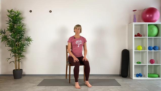 Frau in rosa T-Shirt sitzt barfuß auf einem Stuhl, umgeben von Fitnessutensilien und Pflanzen in einem hellen Raum.