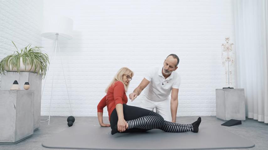 Eine Frau in rot und ein Mann in Weiß demonstrieren eine Gymnastikübung auf einer Matte in einem hellen Raum.