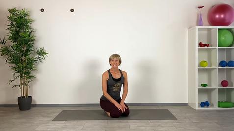 Frau sitzt lächelnd auf einer Yoga-Matte, umgeben von Fitnessgeräten und Pflanzen in einem hellen Raum.