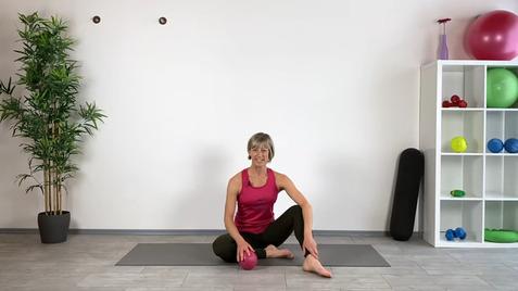 Frau im Sportoutfit sitzt auf einer Matte, hält einen pinken Ball und lächelt, daneben Fitnessgeräte in Regalen.