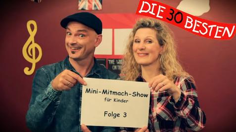 Zwei Personen halten ein Schild mit der Aufschrift „Mini-Mitmach-Show für Kinder, Folge 3“ vor einer bunten Wand.