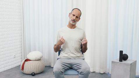 Ein Mann sitzt auf einem Stuhl und zeigt Bewegungen mit den Händen in einem hellen Raum mit Vorhängen.