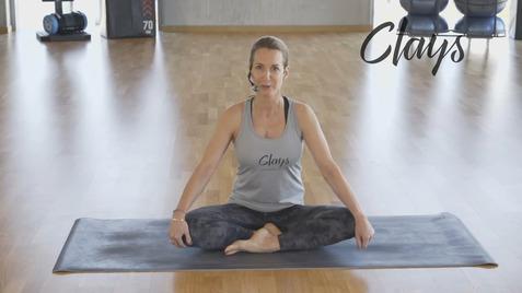 Frau sitzt im Schneidersitz auf einer Matte im Fitnessstudio und blickt in die Kamera, trägt ein sportliches Oberteil.