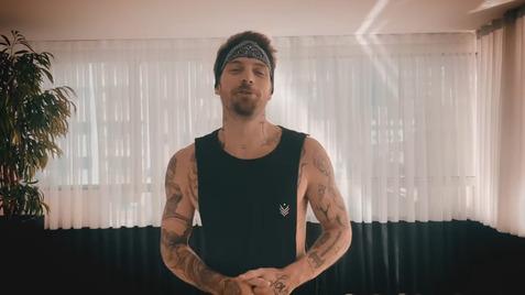 Mann mit Tätowierungen trägt ein schwarzes Tanktop und Bandana, steht in einem hellen Raum mit Pflanzen und Vorhängen.