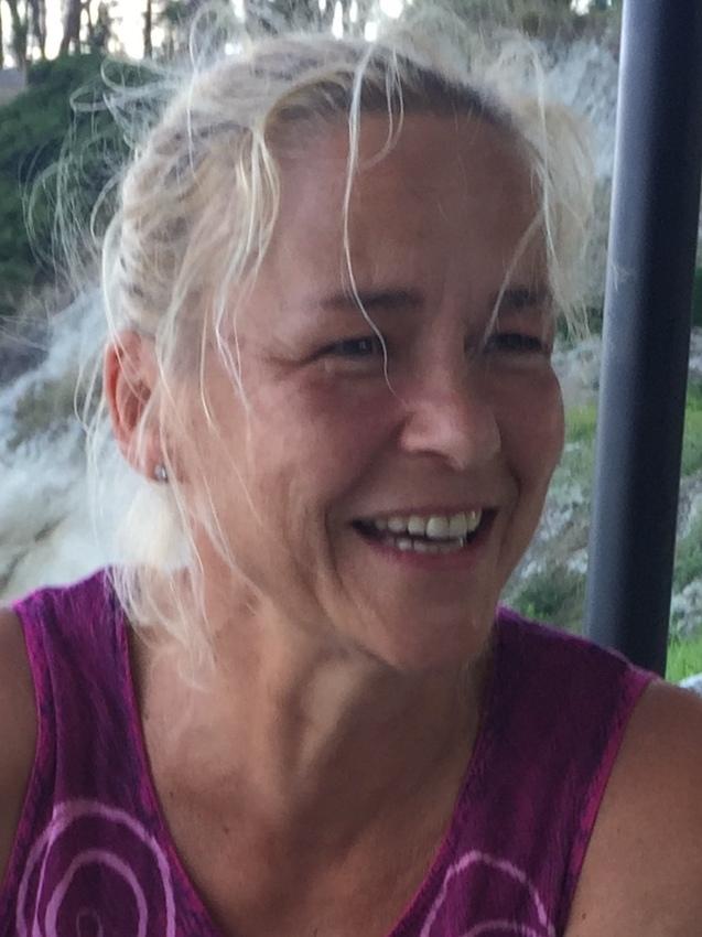 Lächelnde Frau mit blonden, lockigen Haaren, die ein lila Oberteil trägt, vor einer natürlichen Kulisse.