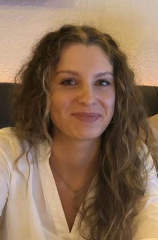 Portrait einer lächelnden Frau mit lockigem, braunem Haar in einem hellen, etwas unscharfen Hintergrund.