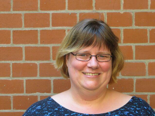 Frau mit blonden, schulterlangen Haaren und Brille lächelt vor einer Ziegelsteinwand. Sie trägt ein dunkelblaues Oberteil.