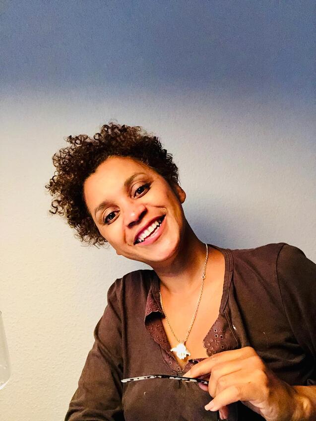 Lächelnde Frau mit lockigem Haar hält eine Schere in der Hand, vor einem neutralen Hintergrund.