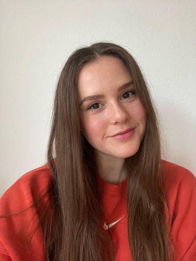 Porträt einer Person mit langen, braunen Haaren, die in einem roten Sweatshirt vor einer weißen Wand lächelt.