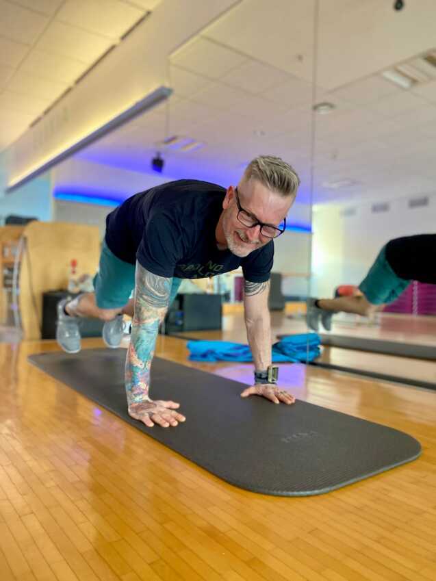 Mann mit Tattoos in Sportkleidung macht ein Plank-Training auf einer Matte in einem Fitnessstudio.
