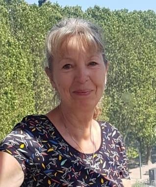 Lächelnde Frau mit blonder, halblanger Haarfrisur, trägt ein gemustertes Oberteil vor grüner Kulisse.