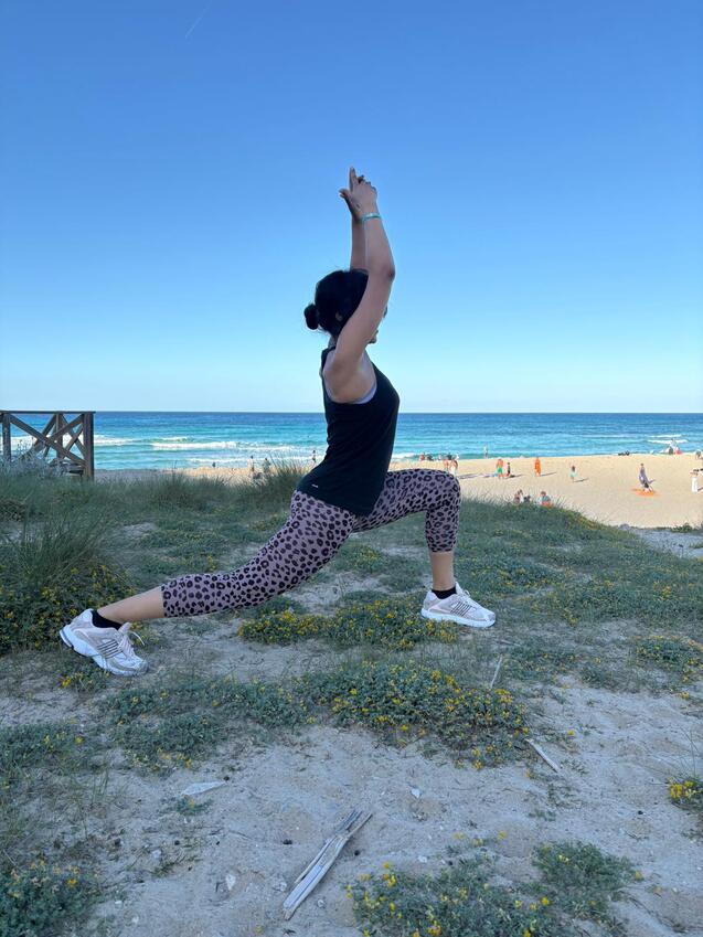Frau in sportlicher Kleidung führt eine Dehnübung am Strand mit Meer im Hintergrund durch.
