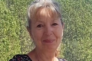 Lächelnde Frau mit blonder, halblanger Haarfrisur, trägt ein gemustertes Oberteil vor grüner Kulisse.