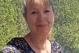 Lächelnde Frau mit blonder, halblanger Haarfrisur, trägt ein gemustertes Oberteil vor grüner Kulisse.