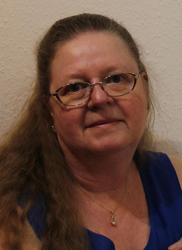 Porträt einer Frau mit langen, lockigen Haaren, Brille und blauer Oberbekleidung vor einer hellen Wand.