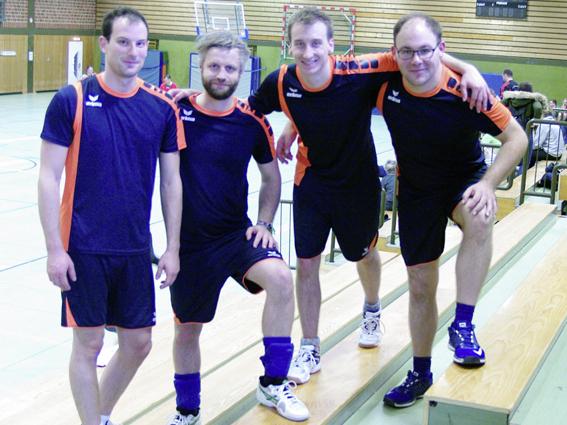 Vier Männer in Sportkleidung posieren auf einer Holztribüne in einer Sporthalle.