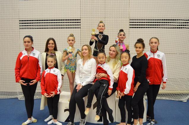 Gruppe von Gymnastinnen in roten Trainingsanzügen posiert um eine Gewinnerin mit Pokalen auf einem Treppchen.