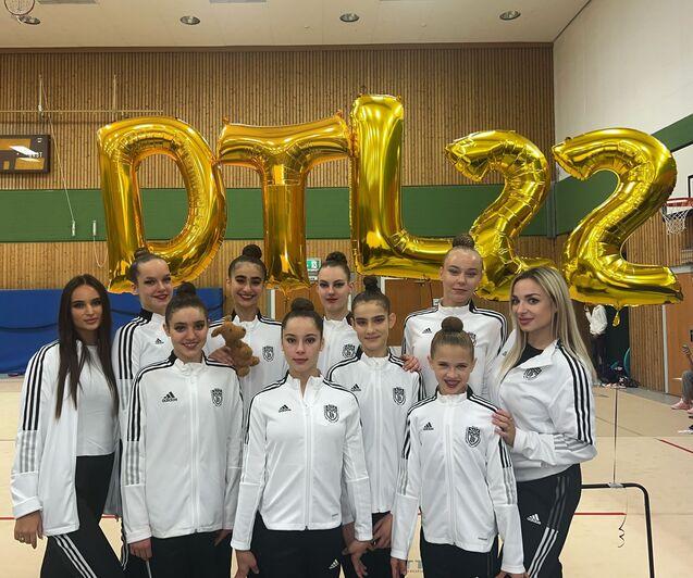 Gruppe von Tänzerinnen in weißen Trainingsjacken, posierend vor großen goldenen Ballons mit der Aufschrift „DTL22“.