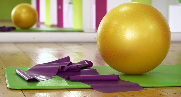 Goldener Fitnessball und violette Stretchbänder auf einer grünen Yogamatte in einem hellen Trainingsraum.