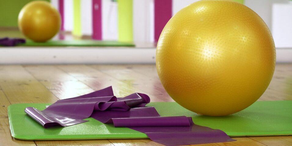 Goldener Fitnessball und violette Stretchbänder auf einer grünen Yogamatte in einem hellen Trainingsraum.