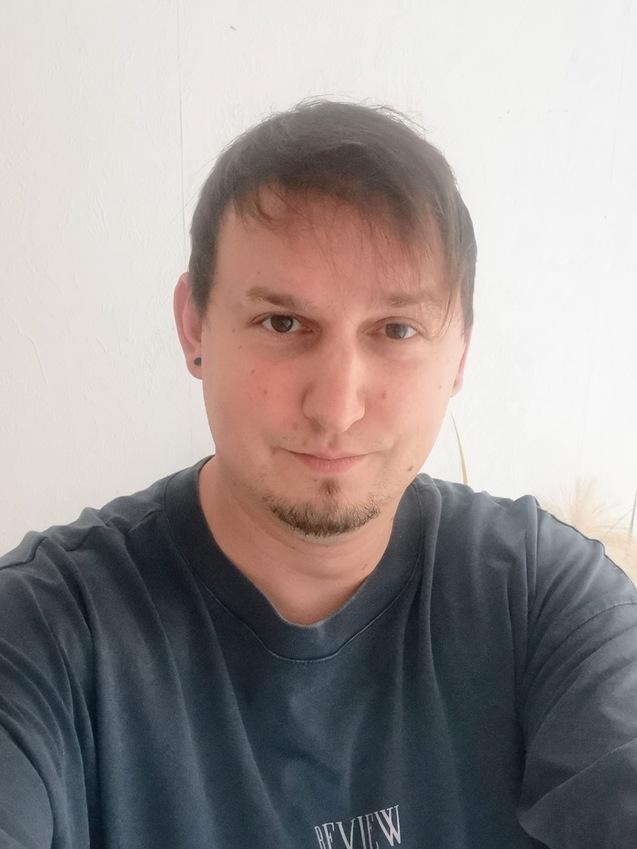 Nahaufnahme eines Mannes mit Kurzhaarfrisur, der ein graues T-Shirt mit Aufschrift trägt, vor einer weißen Wand.