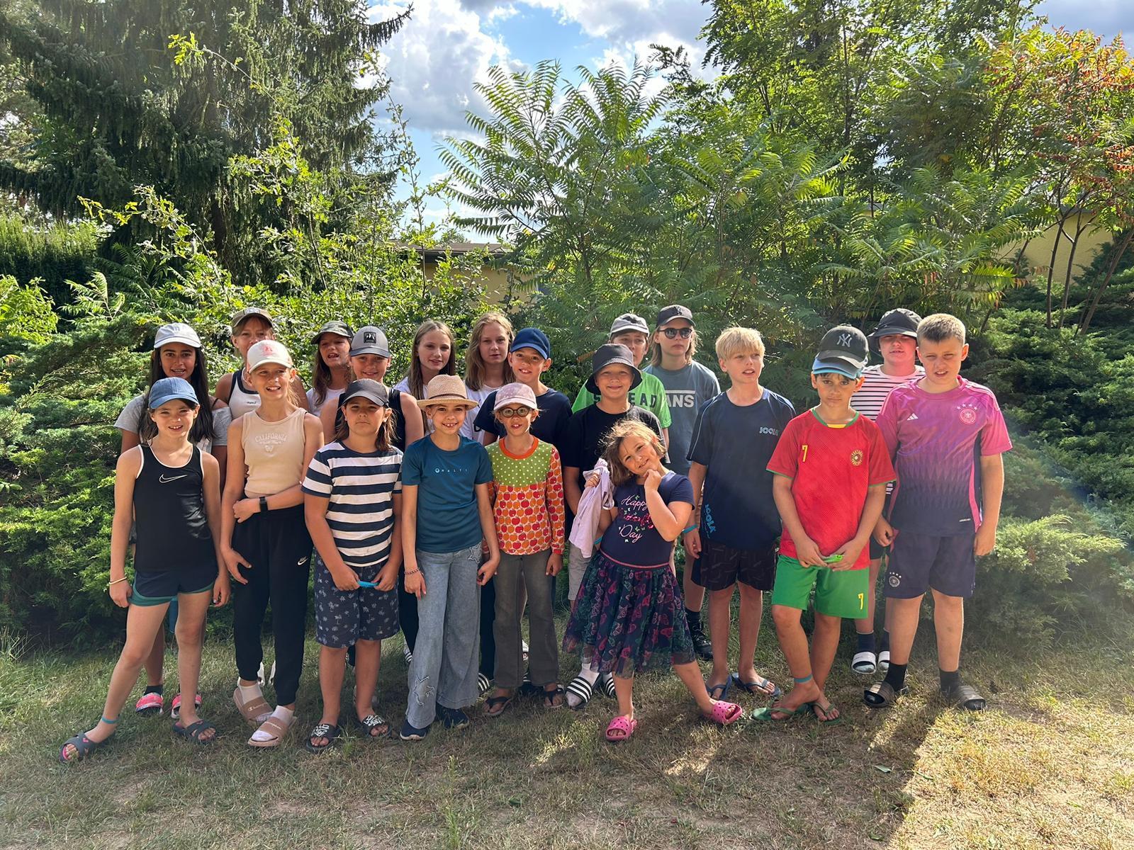 Gruppe von 20 Kindern steht vor Büschen, die meisten tragen Hüte, sonnige Stimmung, verschiedene Kleidungsstile.